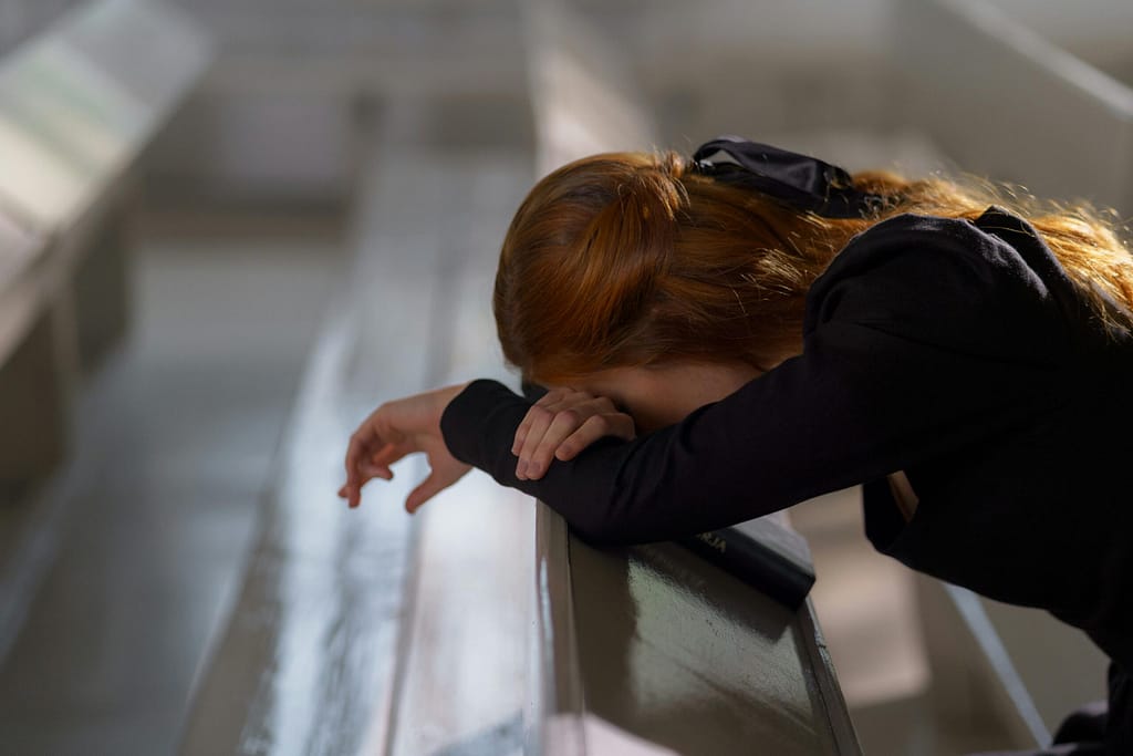 A redheaded woman in deep prayer, leaning on a church pew in a serene atmosphere.