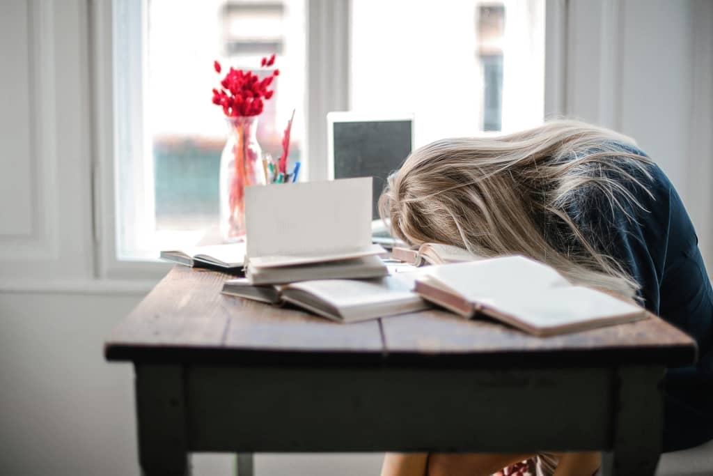 Starting Over After Failure Blonde woman slumped over desk filled with books, showcasing study fatigue indoors.