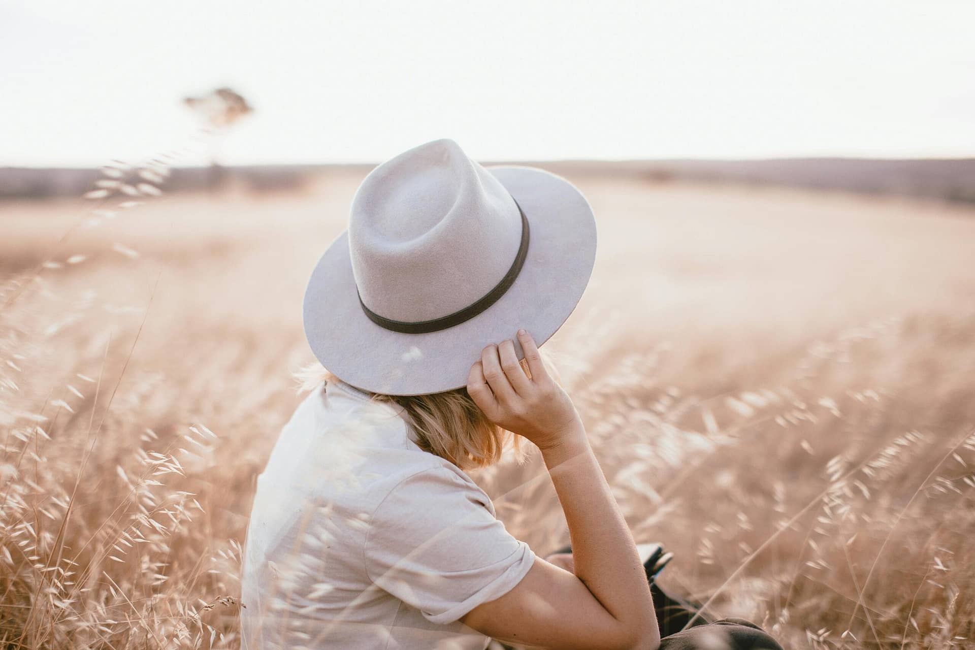 A woman with a gray hat sits in a serene wheat field under natural light.